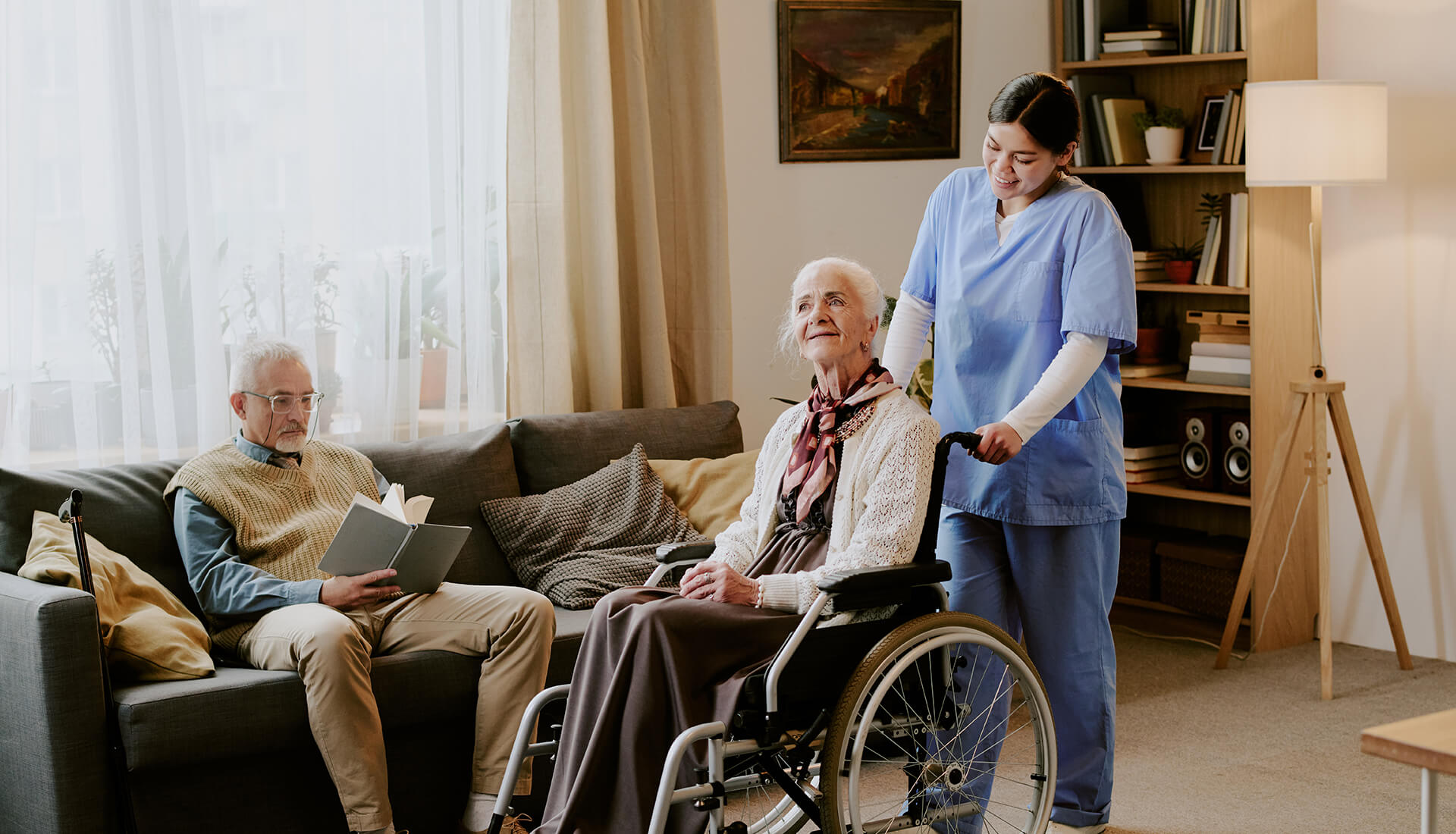 Caregiver assisting an older woman in a wheelchair at home while a man reads on the sofa